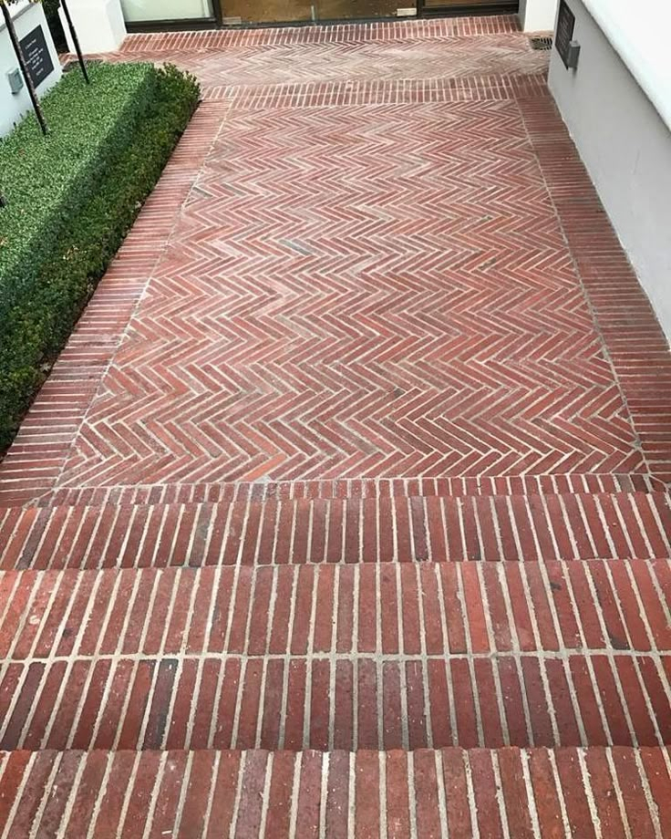 Classic red herringbone-patterned brick front entry paving with contrasting straight border course, boxwood hedge to the side, adjacent to white exterior wall.