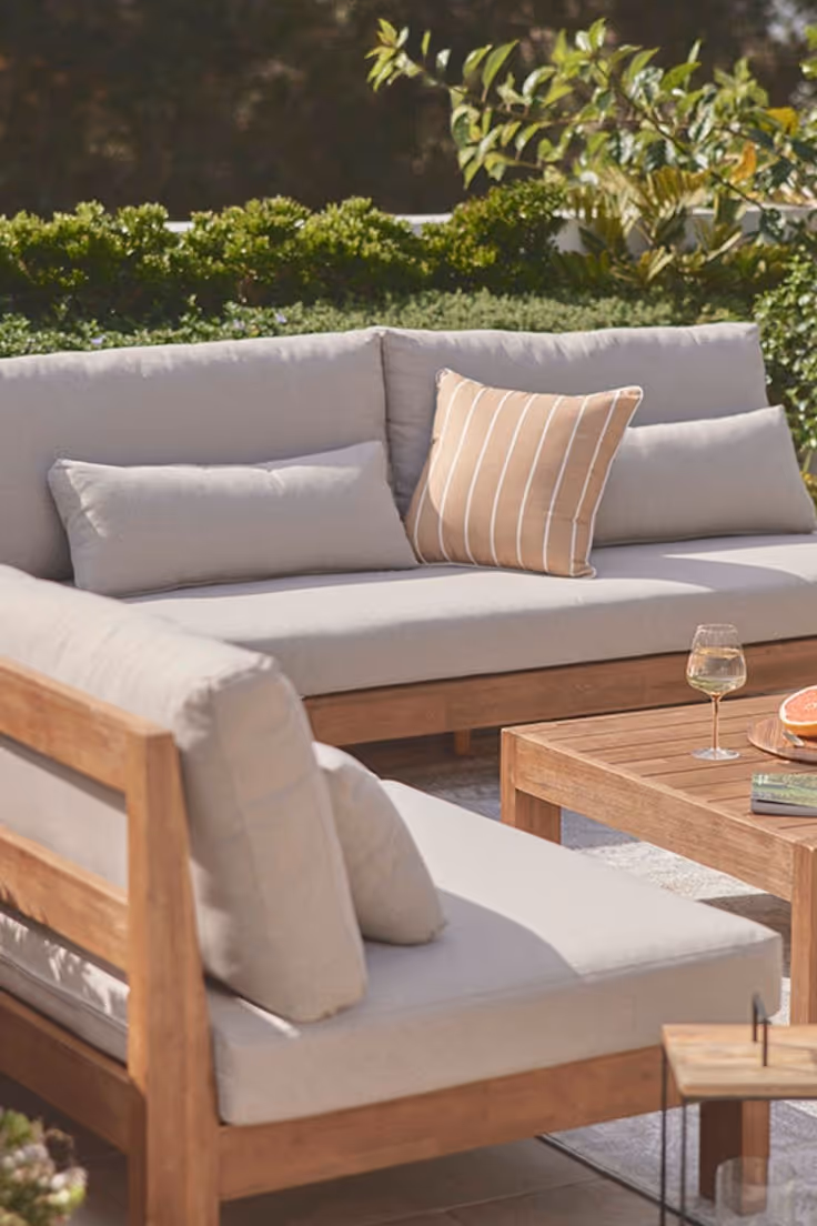 Close-up of a natural teak outdoor sectional sofa with light gray cushions, striped accent pillow, teak coffee table with wine glass and grapefruit, with green hedges in background.