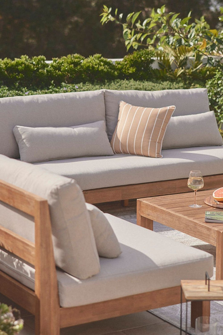 Close-up of a natural teak outdoor sectional sofa with light gray cushions, striped accent pillow, teak coffee table with wine glass and grapefruit, with green hedges in background.