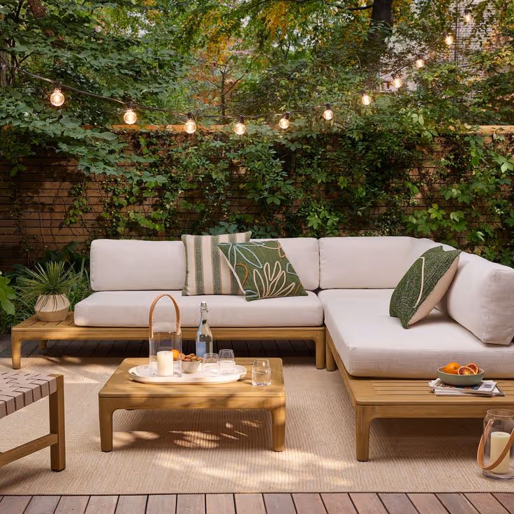 Backyard wood deck with teak sectional sofa, cream and green leaf-print cushions, square coffee table with tray, string lights overhead, and lush green vine-covered fence behind.