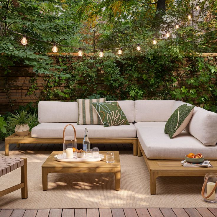 Backyard wood deck with teak sectional sofa, cream and green leaf-print cushions, square coffee table with tray, string lights overhead, and lush green vine-covered fence behind.