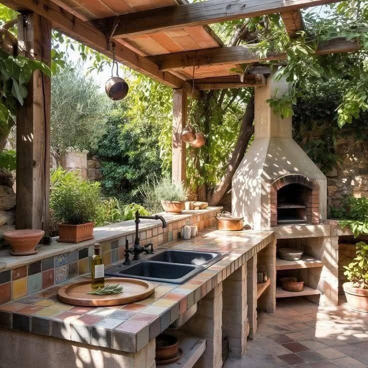 Rustic Mediterranean outdoor kitchen under a wood pergola featuring a brick pizza oven, colorful hand-painted tile countertop, sink with copper pots, potted herbs, and surrounding garden.