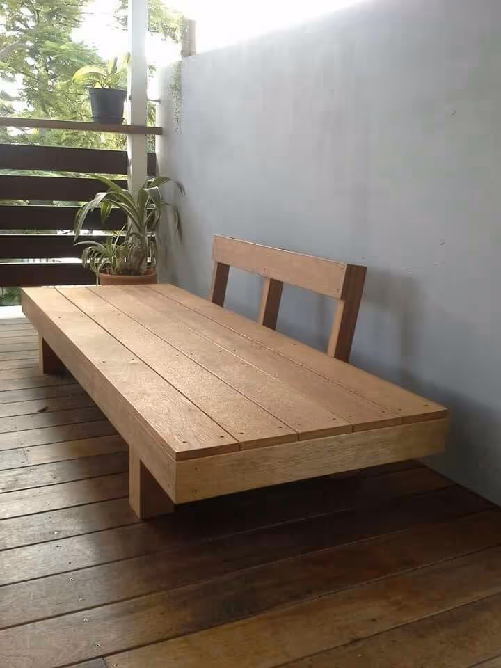 Simple DIY wood platform daybed on a covered deck with horizontal slat railing, potted plants, and tropical trees visible beyond the balcony.