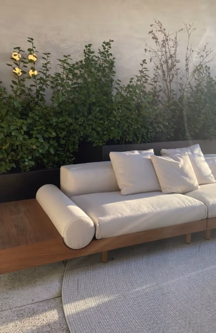 Minimalist outdoor patio with a cream cushion teak sofa on a round woven rug, positioned in front of a long black metal raised planter filled with leafy shrubs on a concrete surface.