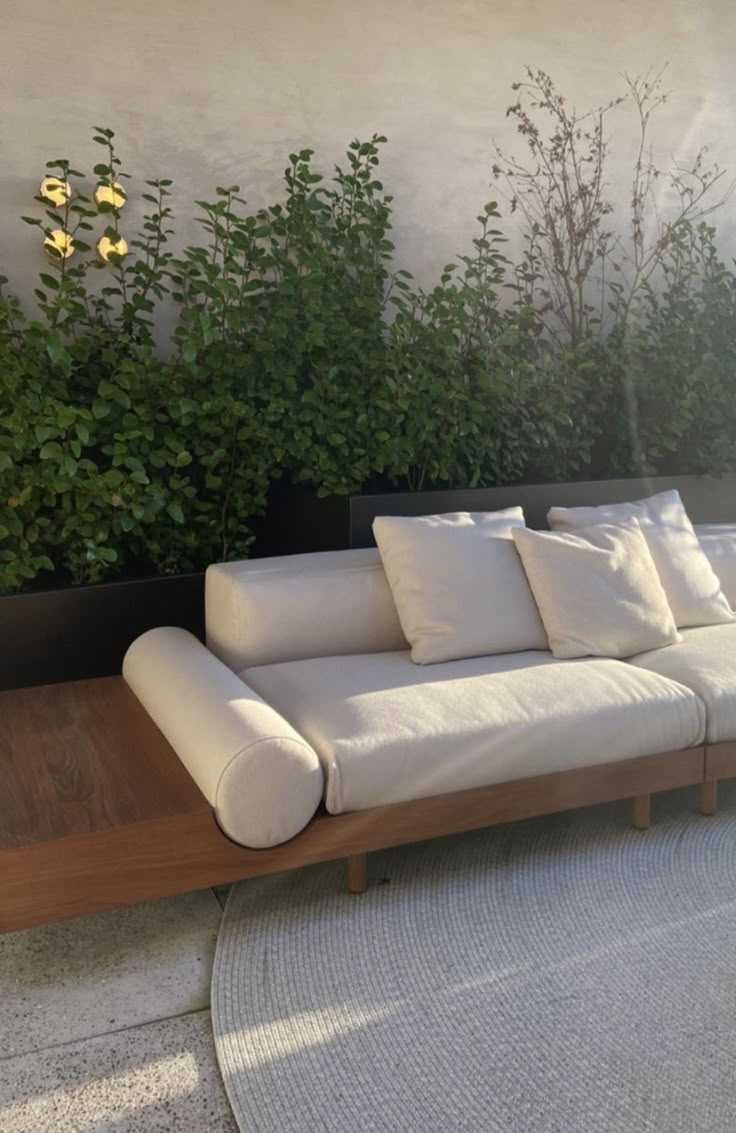 Minimalist outdoor patio with a cream cushion teak sofa on a round woven rug, positioned in front of a long black metal raised planter filled with leafy shrubs on a concrete surface.