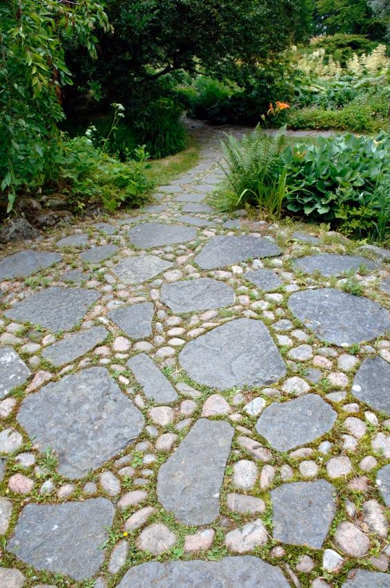 Winding garden path made of irregular grey flagstone pieces set among rounded river pebbles, bordered by ferns, hostas, and lush green woodland plantings under a tree canopy.