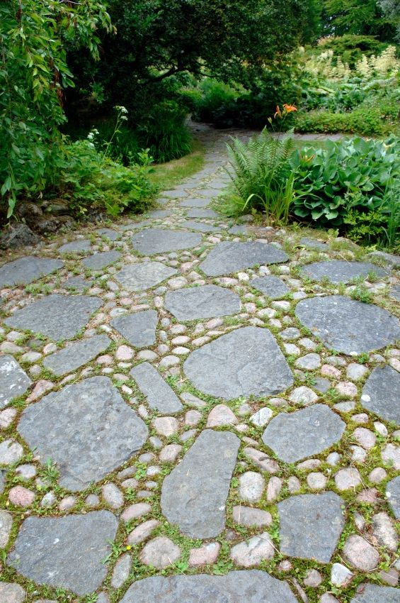 Winding garden path made of irregular grey flagstone pieces set among rounded river pebbles, bordered by ferns, hostas, and lush green woodland plantings under a tree canopy.
