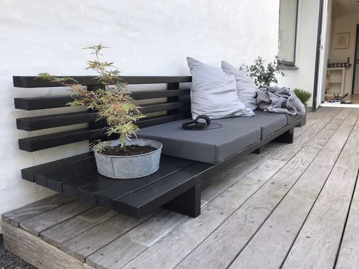 Minimalist black-stained wood outdoor bench with gray cushion and white pillow, a potted Japanese maple beside it, on a weathered wood deck next to a white stucco wall.