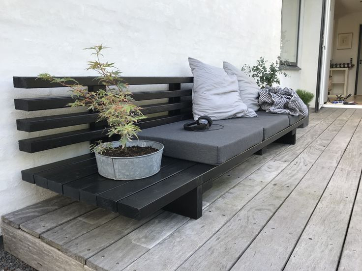 Minimalist black-stained wood outdoor bench with gray cushion and white pillow, a potted Japanese maple beside it, on a weathered wood deck next to a white stucco wall.