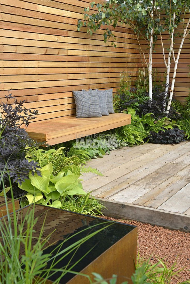 Modern garden with horizontal cedar slat privacy wall, floating teak bench with gray cushions, corten steel water basin, birch trees, ferns, hostas, and black mulch planting beds.