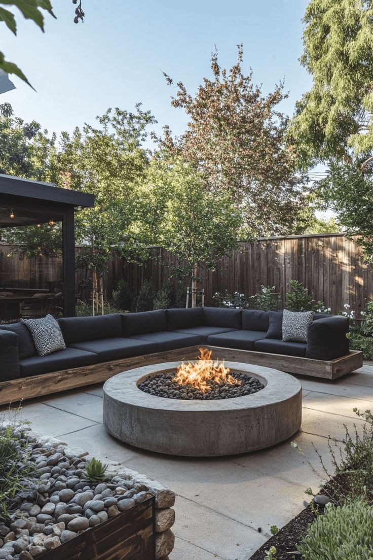 Modern suburban backyard with a large round concrete fire pit bowl, built-in wood frame sectional bench with dark cushions on a concrete paver patio, surrounded by mature trees and fence.