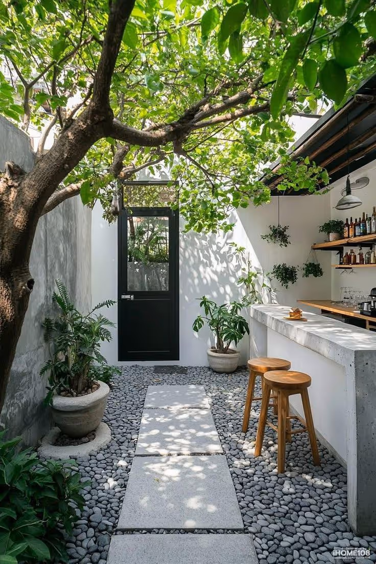 Narrow enclosed courtyard with a concrete bar counter and wood stools, stepping stone path through river pebbles, mature shade tree, potted tropical plants, and a wall-mounted bar shelf with plants.