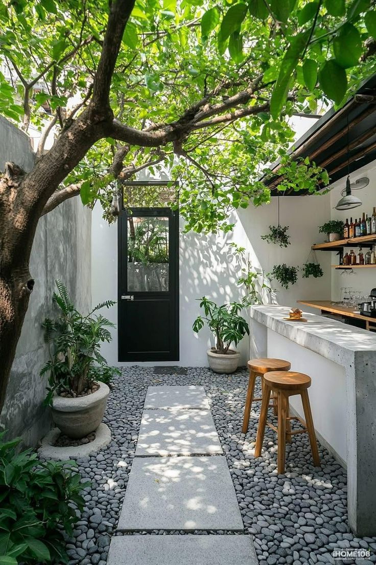 Narrow enclosed courtyard with a concrete bar counter and wood stools, stepping stone path through river pebbles, mature shade tree, potted tropical plants, and a wall-mounted bar shelf with plants.