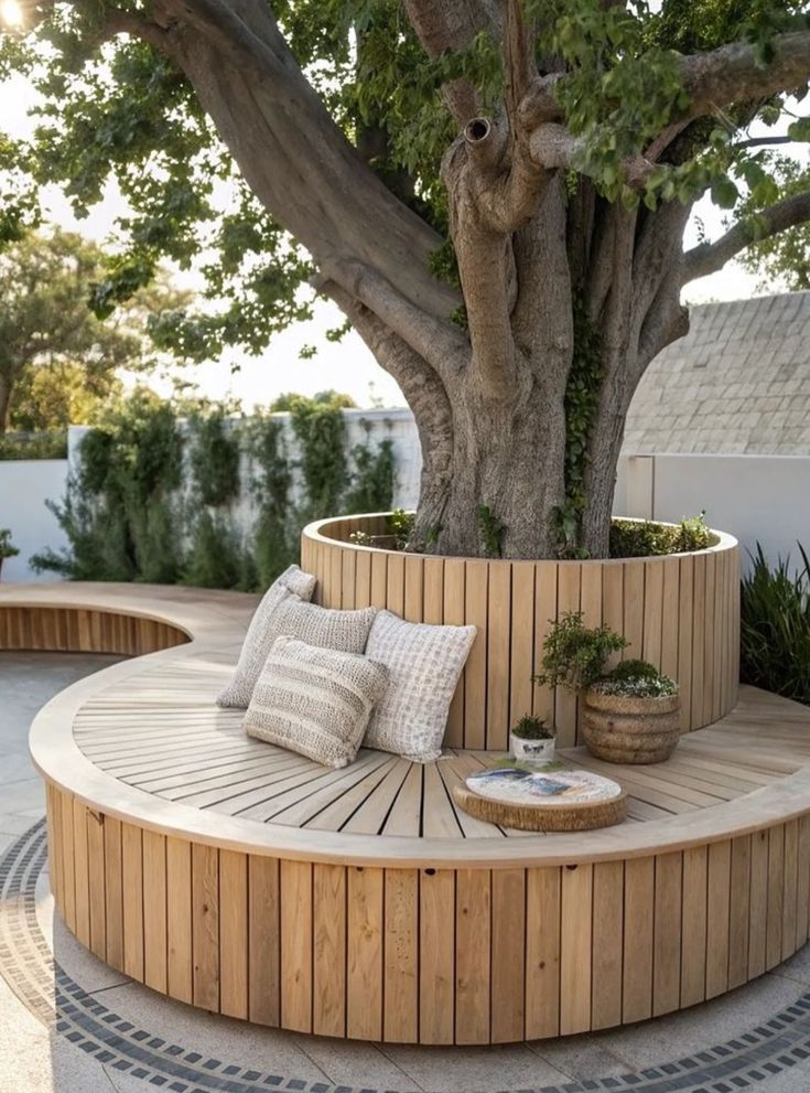 Large circular teak wood built-in bench wrapping around a mature shade tree trunk, with woven cushion pillows, on a decorative stone mosaic patio with white stucco wall backdrop.