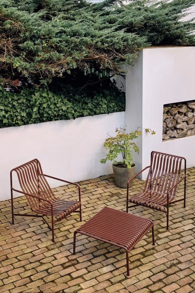 Minimalist walled courtyard with herringbone brick pavers, two terracotta-red metal slatted lounge chairs, a low matching footrest, potted Japanese maple, and a firewood niche in a white stucco wall.