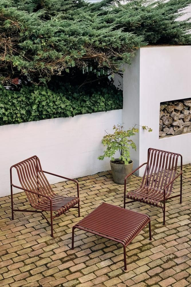 Minimalist walled courtyard with herringbone brick pavers, two terracotta-red metal slatted lounge chairs, a low matching footrest, potted Japanese maple, and a firewood niche in a white stucco wall.