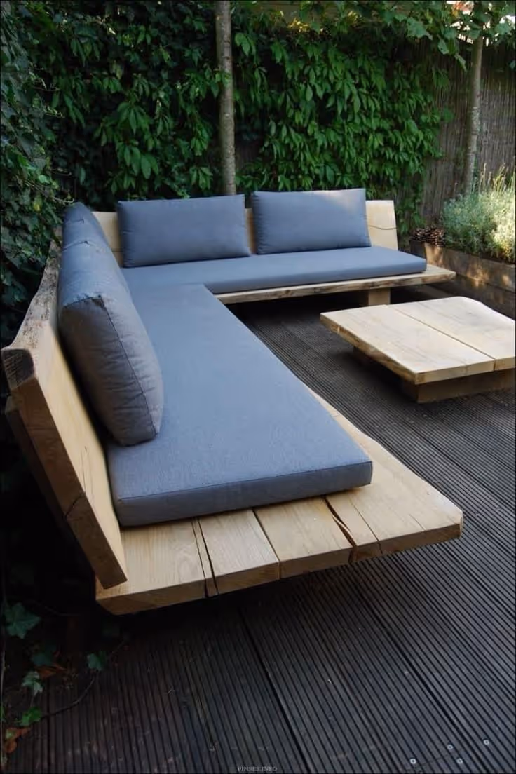 Modern outdoor sectional sofa made from thick natural timber with blue-gray cushions on a dark hardwood deck, backed by a lush green climbing vine privacy wall.