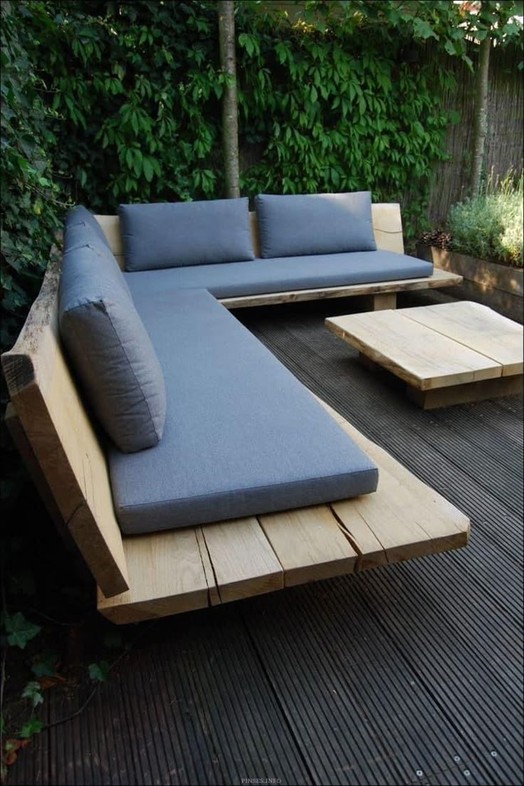 Modern outdoor sectional sofa made from thick natural timber with blue-gray cushions on a dark hardwood deck, backed by a lush green climbing vine privacy wall.