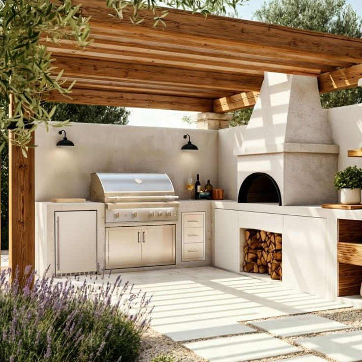 Mediterranean outdoor kitchen under a cedar pergola with stucco walls, stainless steel built-in grill, arched wood-fired pizza oven, firewood storage, and lavender plants in foreground.