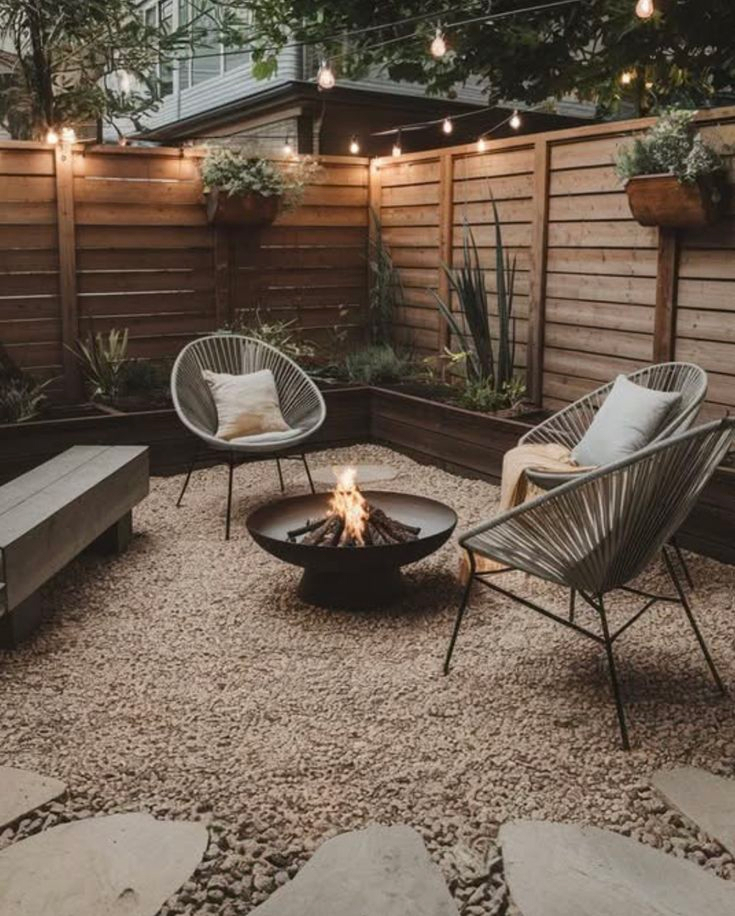 Small urban backyard with decomposed granite ground cover, black steel fire bowl, two gray Acapulco lounge chairs, raised planter beds along horizontal wood fence, and string lights overhead.