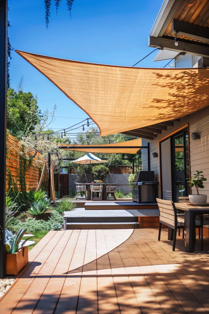 Contemporary backyard with a two-level hardwood deck, large triangular tan shade sail, outdoor dining set, and a second shade umbrella in a lower landscaped garden area with native shrubs and agave plants.