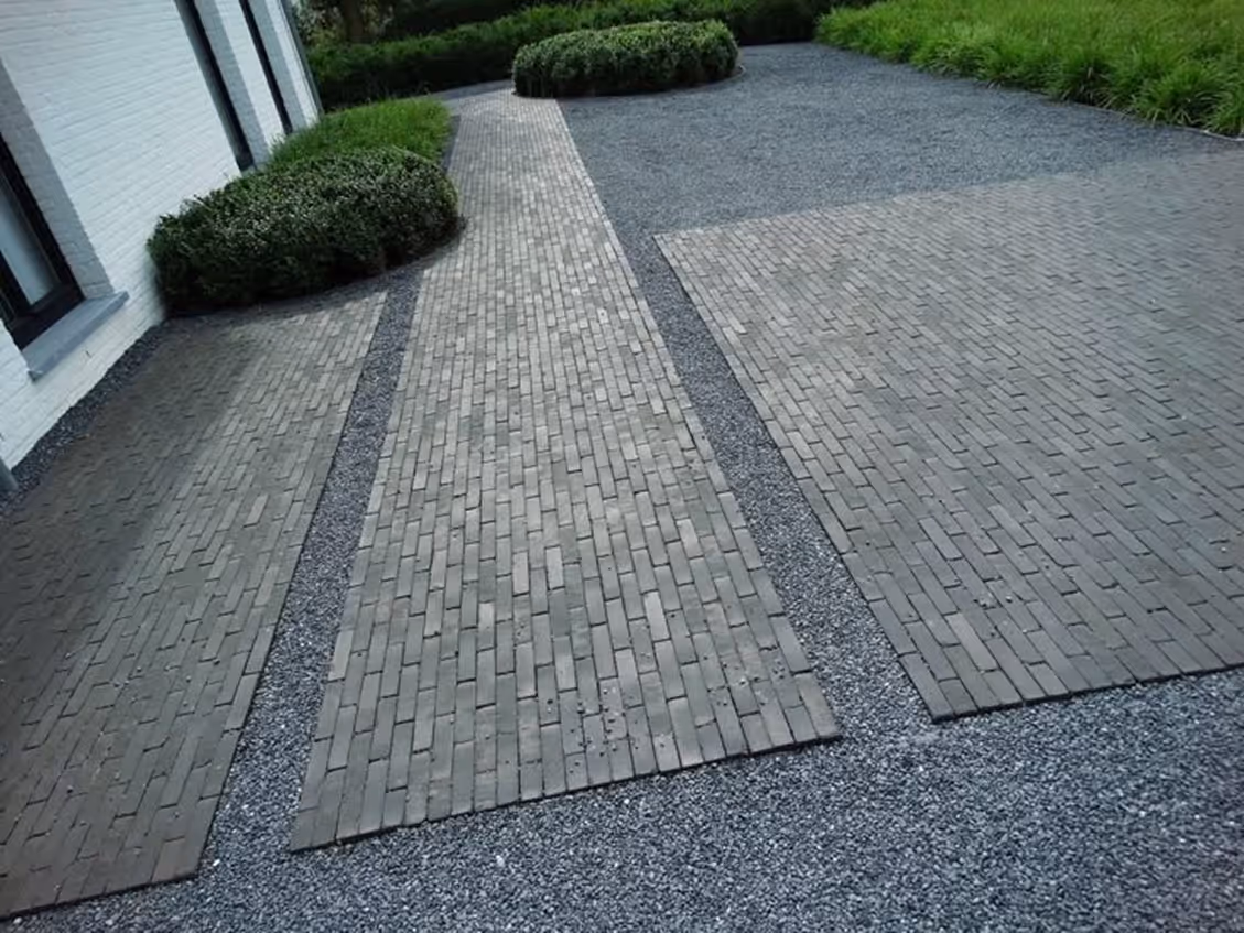 Modern front yard with dark gray gravel ground cover, dark brick paver pathway and parking pad sections, trimmed boxwood hedges, and ornamental grasses along a white modern home facade.
