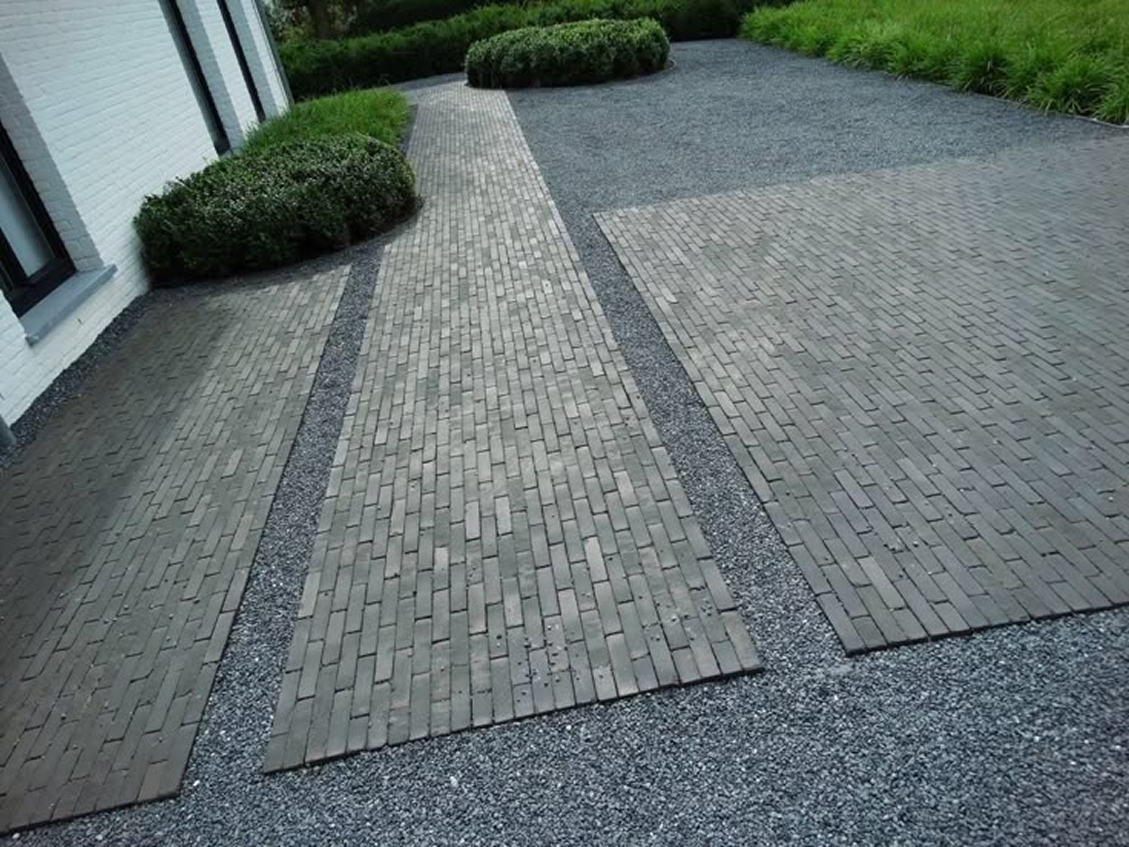 Modern front yard with dark gray gravel ground cover, dark brick paver pathway and parking pad sections, trimmed boxwood hedges, and ornamental grasses along a white modern home facade.