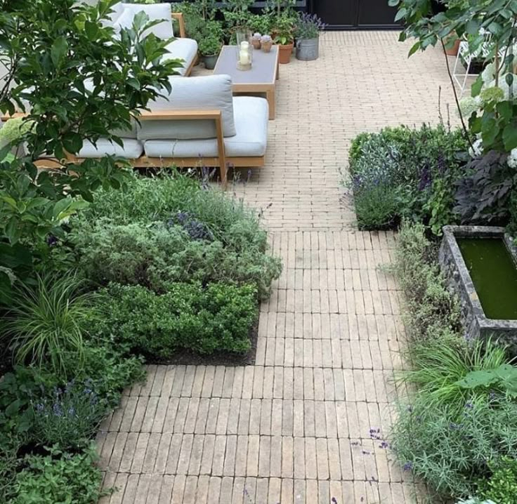 An aerial view of a structured garden with a long brick paver pathway leading to an outdoor seating area with a teak sectional and coffee table, flanked by lush planting beds and a raised stone water feature trough.