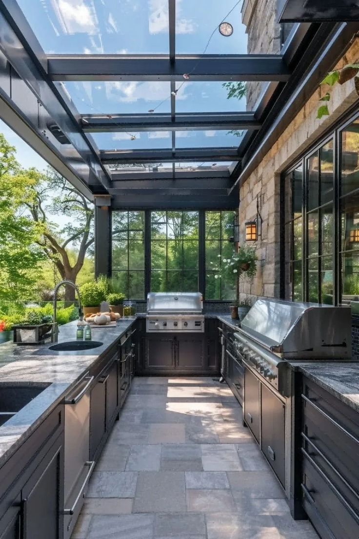 A high-end enclosed outdoor kitchen with a glass and steel roof structure, dark cabinet countertops, dual stainless steel grills, and black-framed floor-to-ceiling windows overlooking a lush green garden.