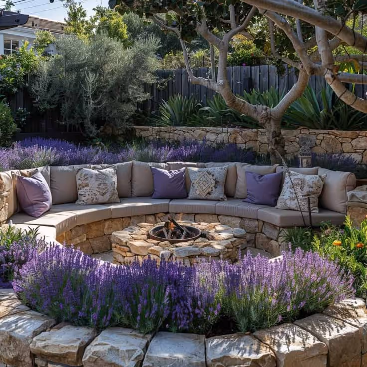 A curved built-in stone bench with cushions and decorative pillows encircles a small fire pit, surrounded by blooming lavender, ornamental grasses, agave, olive trees, and a stone retaining wall.