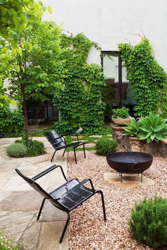 A minimal urban courtyard with large flagstone pavers, gravel ground cover, a cast iron fire bowl on a stand, two black iron lounge chairs, agave plants, and climbing Boston ivy on a white stucco wall.