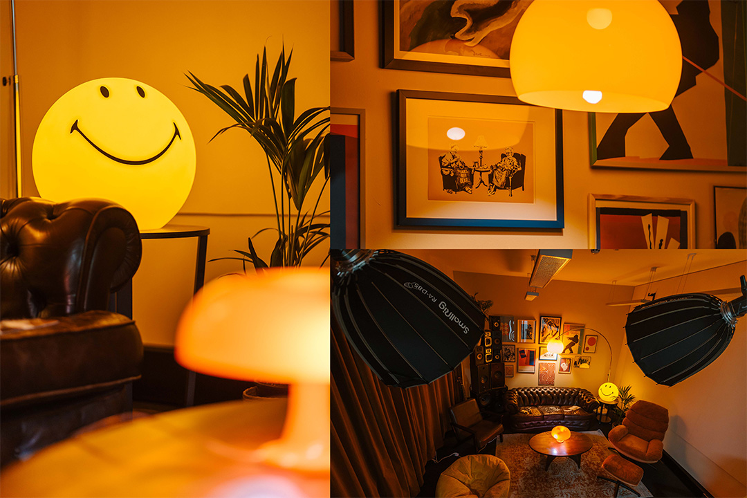Cozy living room vibe studio space with brown leather chairs, a smiley face lamp, round wooden coffee table, and framed art on yellow-lit walls.