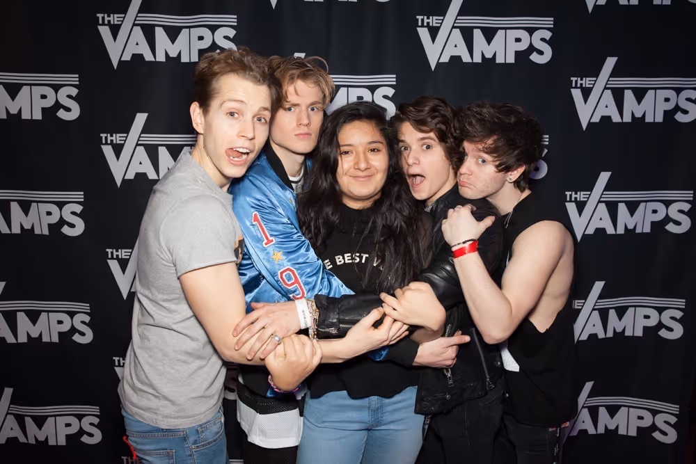 Four members of The Vamps band hugging Aysha in front of a black backdrop with The Vamps logo.