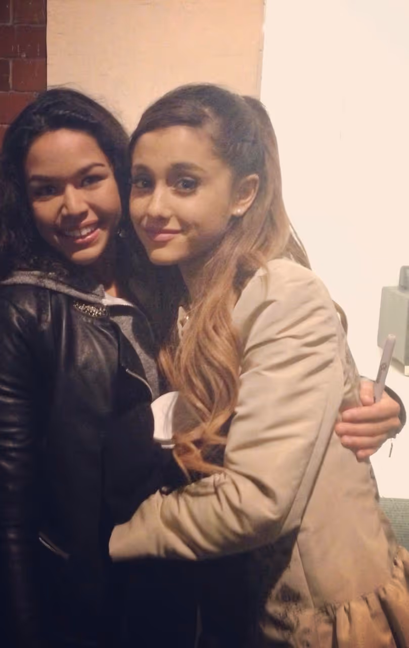 A younger Lauren with Ariana Grande, smiling and hugging. Lauren is wearing a black leather jacket and the Ariana a beige jacket holding a pen.