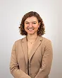 Smiling woman with curly hair wearing a beige blazer over a tan top standing against a white background.