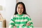 Smiling woman with long brown hair wearing a green and white striped sweater, sitting with arms crossed in a bright room.