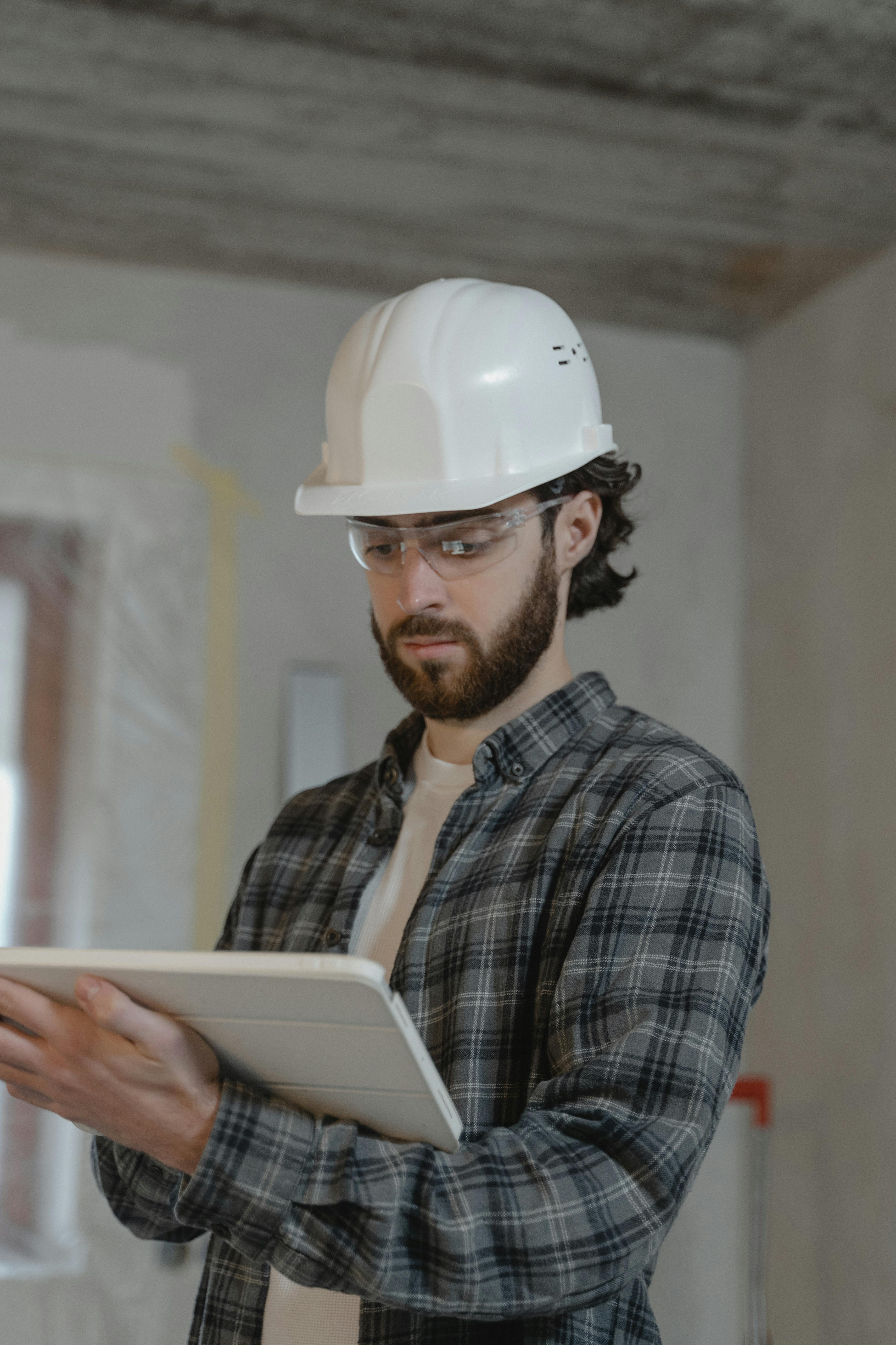 Man wearing a white hard hat and safety glasses, looking at a tablet inside a construction site.