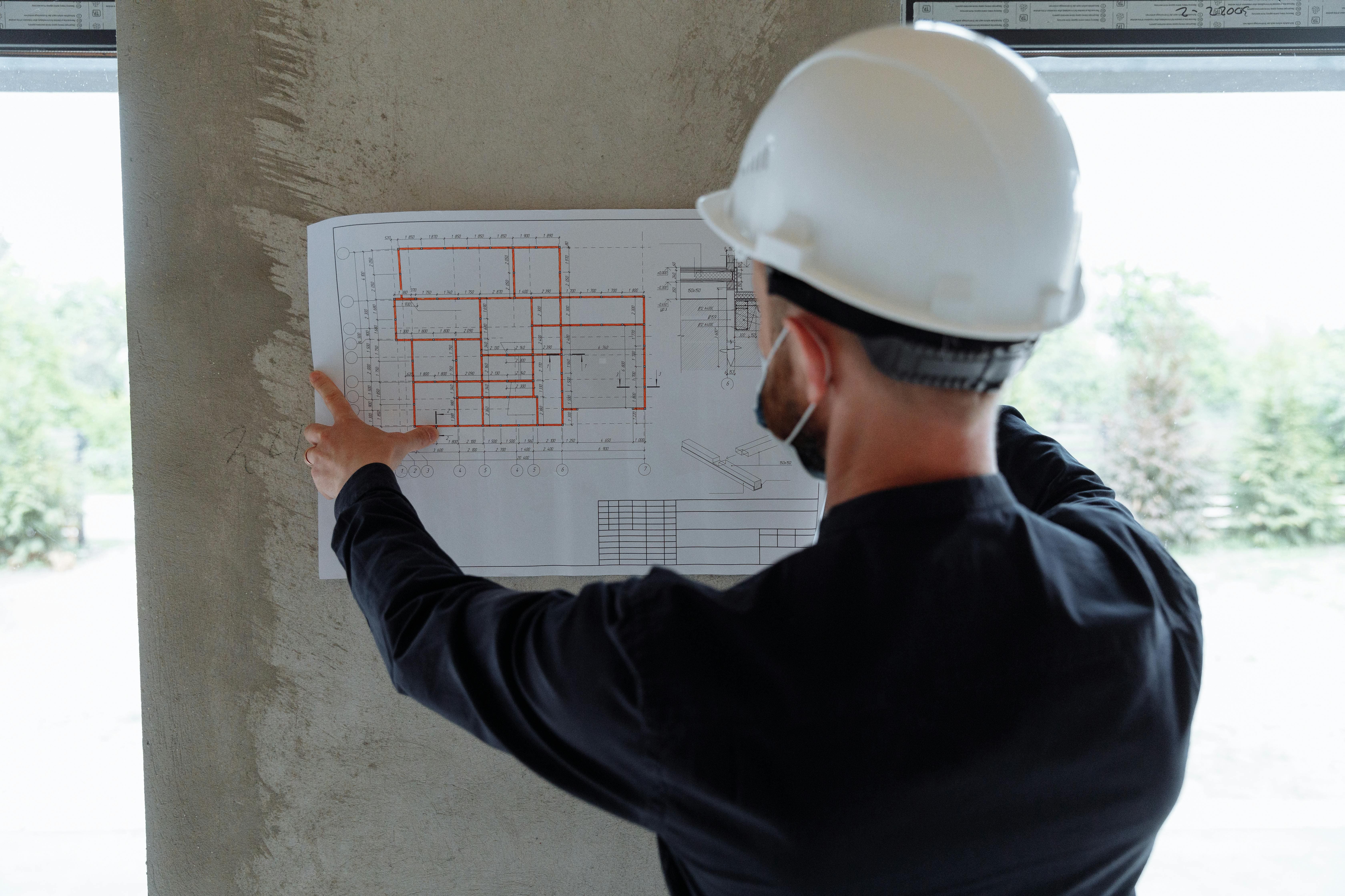 Construction worker wearing a white hard hat and face mask examining a building blueprint with red markings on a concrete wall.