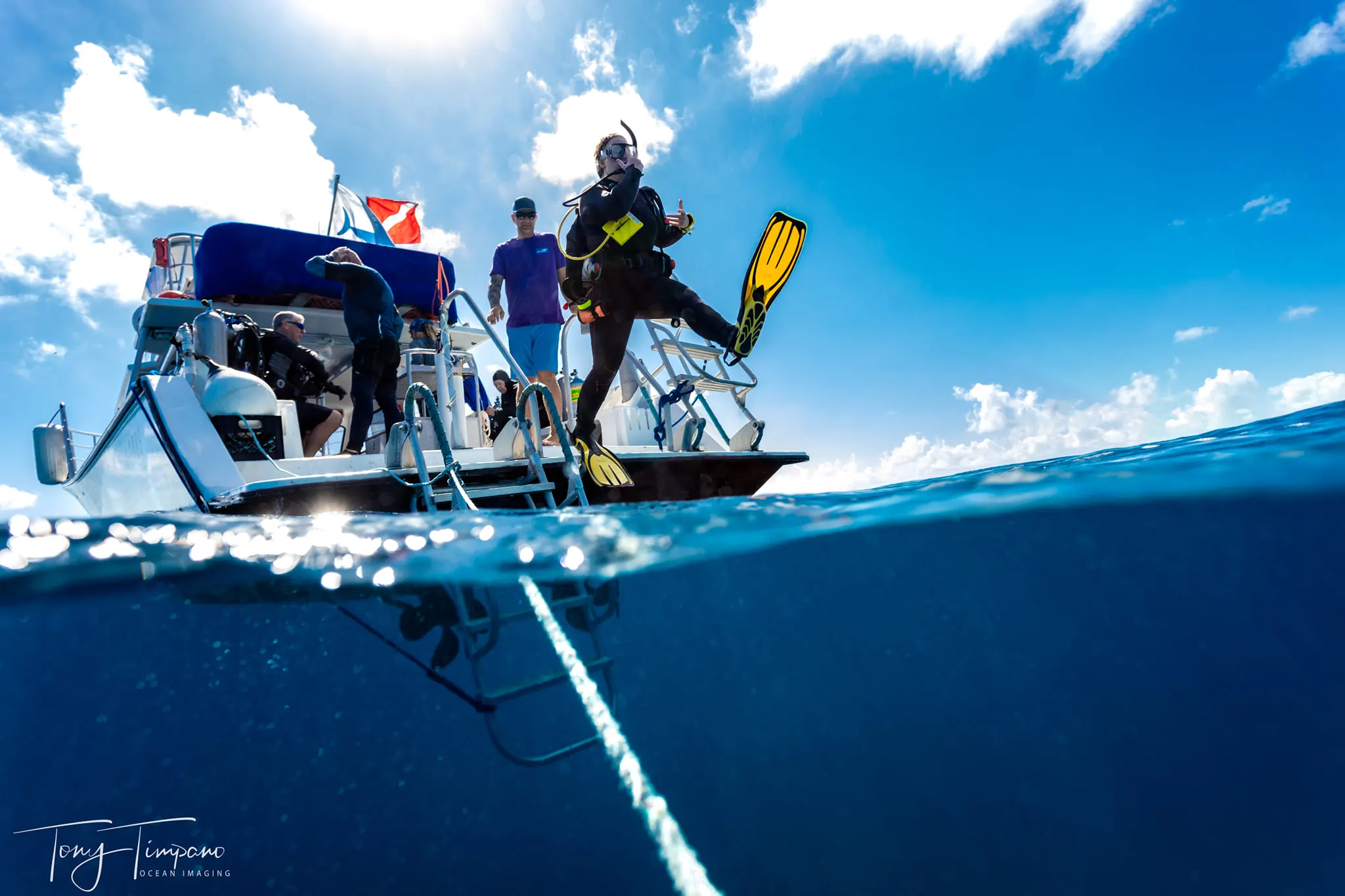 Scuba diver with yellow fins performing a giant stride entry from a boat into clear blue ocean water under a sunny sky.