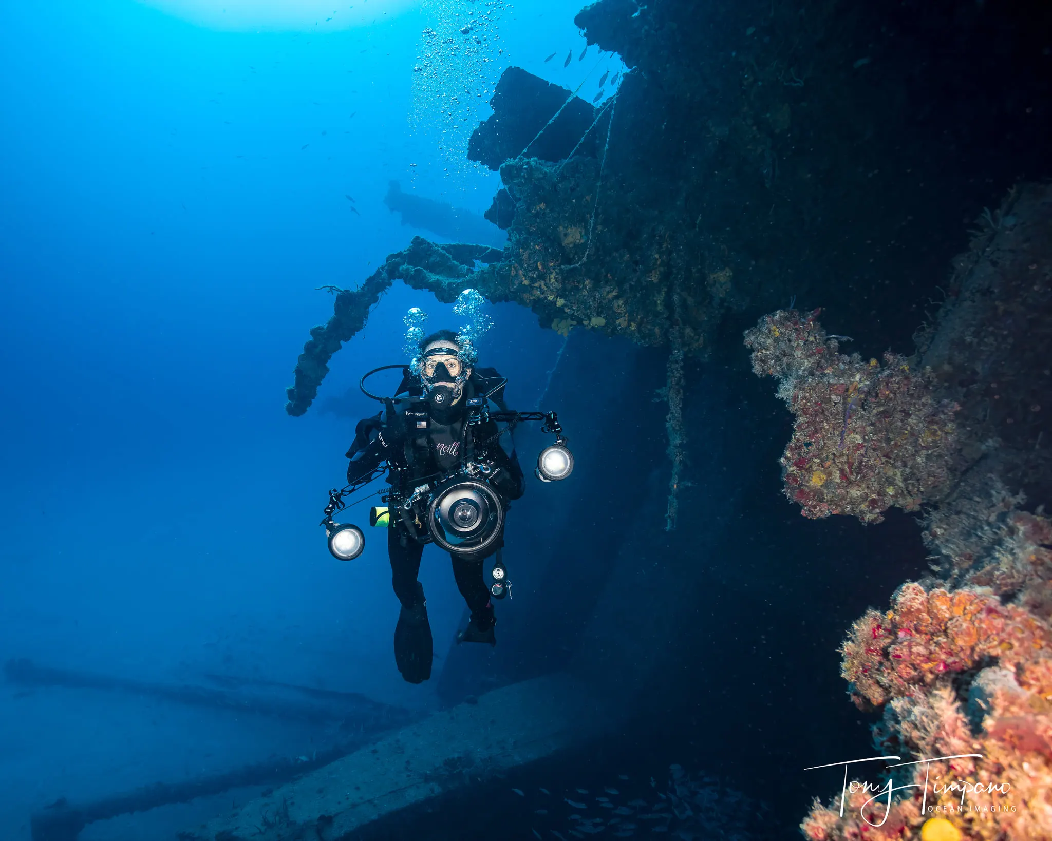 Scuba diver with underwater camera and lights exploring a coral-covered shipwreck in clear blue ocean water.