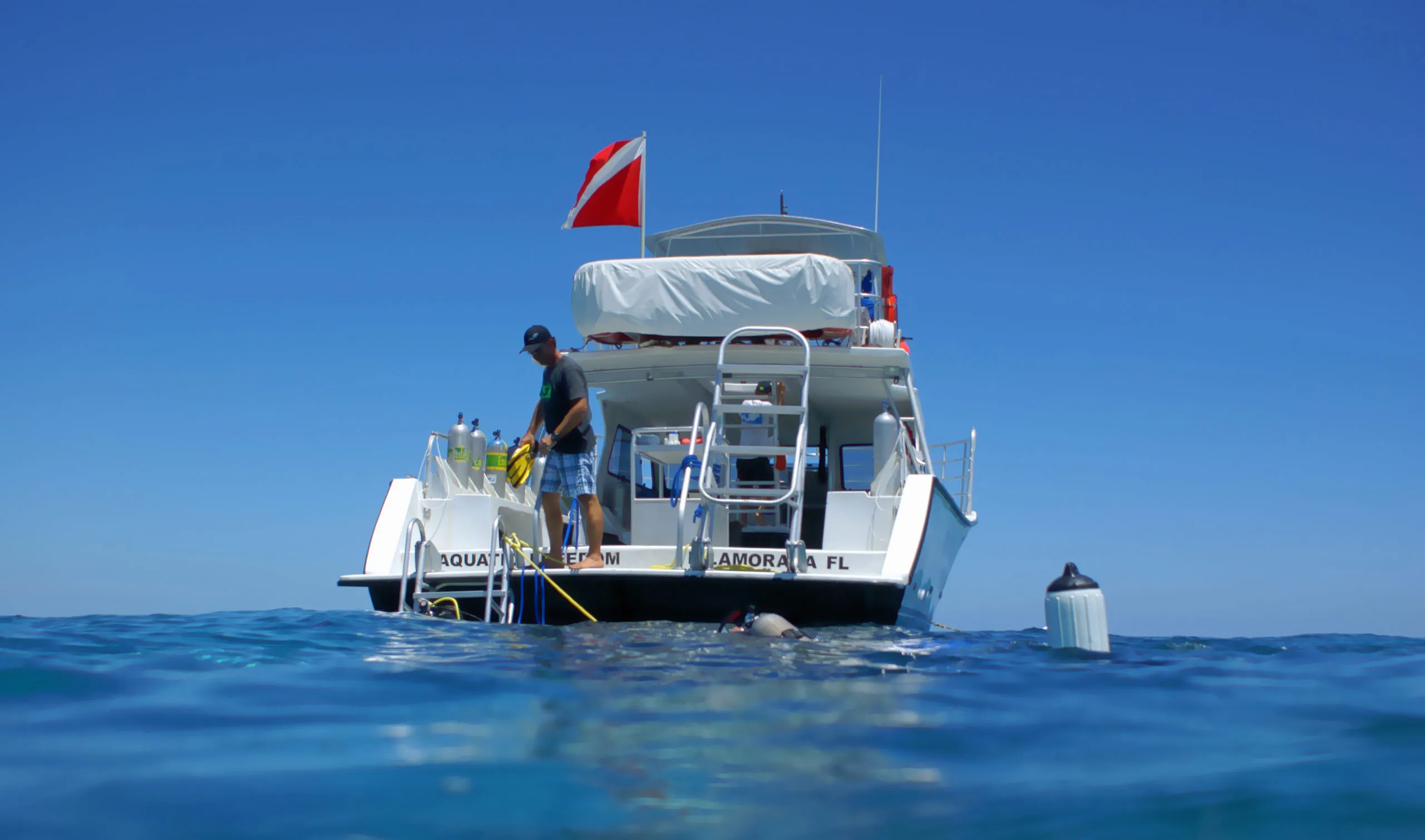 Person standing on the back of an IDC dive boat with scuba tanks and a diver swimming in the ocean.