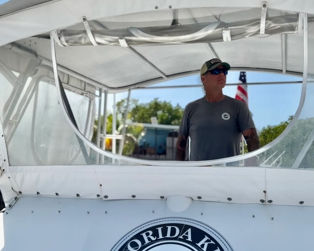 Person wearing sunglasses and a cap standing on a boat with an American flag in the background.