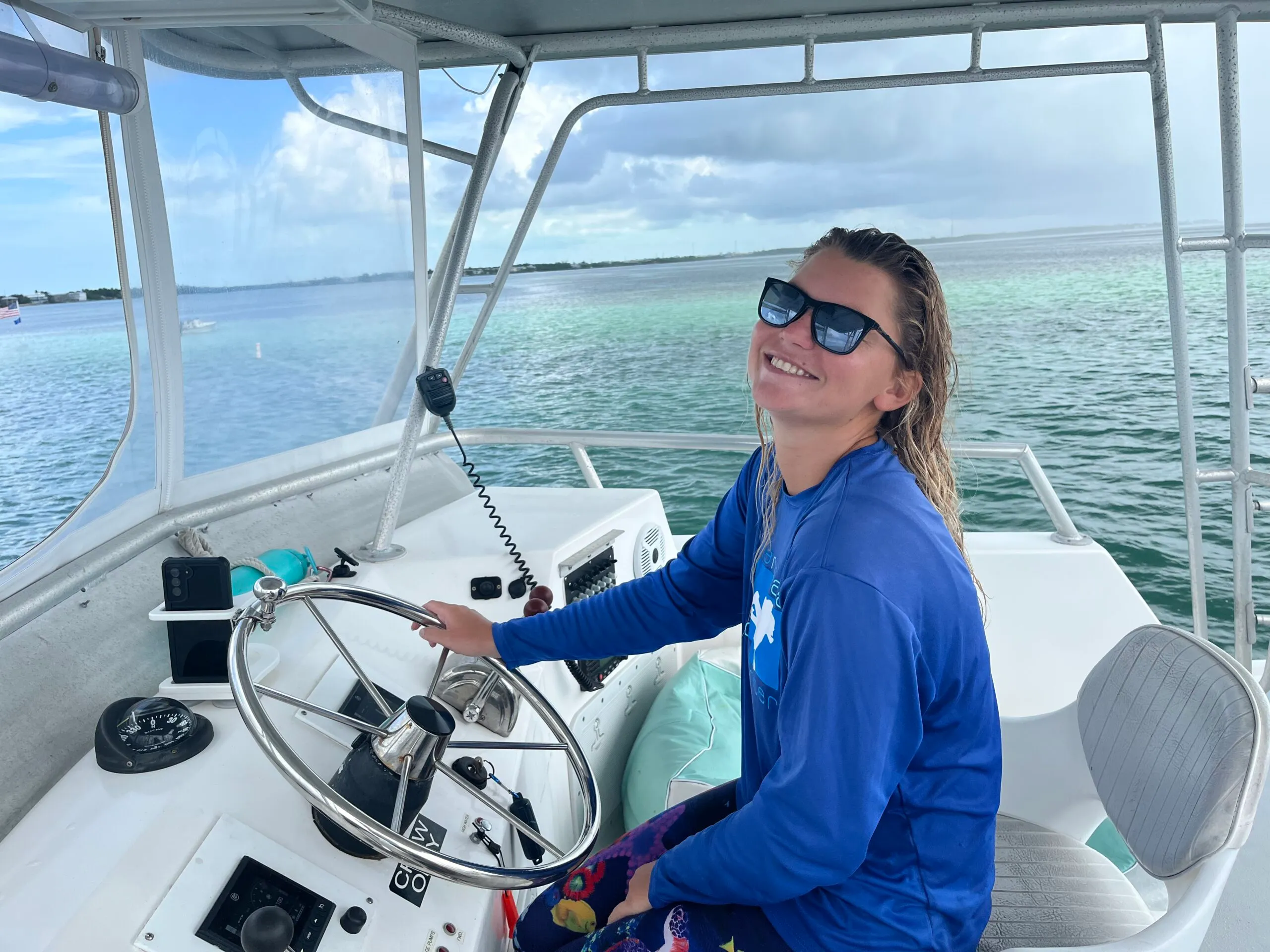 Smiling woman wearing sunglasses and a blue long-sleeve shirt steering a boat with water and sky in the background.