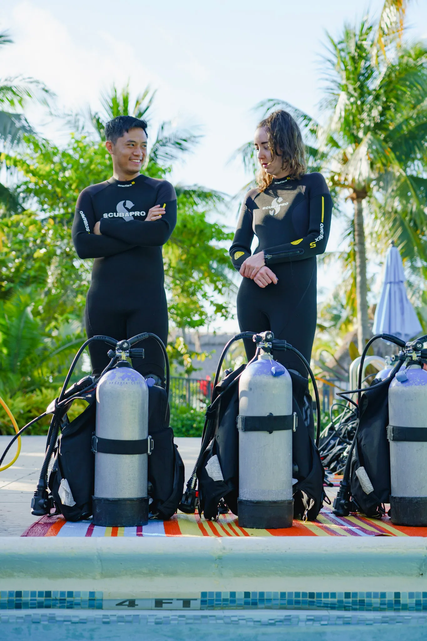 Two scuba divers in black wetsuits standing behind scuba tanks at a poolside with palm trees in the background.