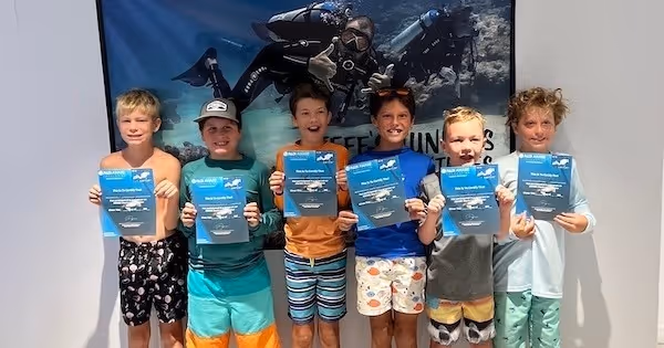 Six kids standing in front of a scuba diving banner, each holding a scuba diving certification card.