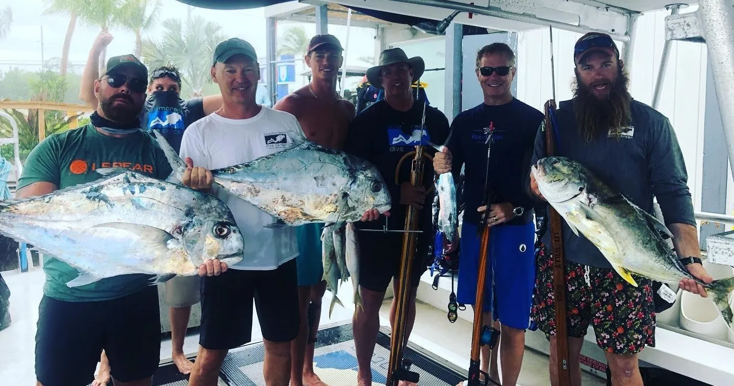 Group of seven men on a boat holding large fish and spearguns, celebrating a successful underwater hunting trip.