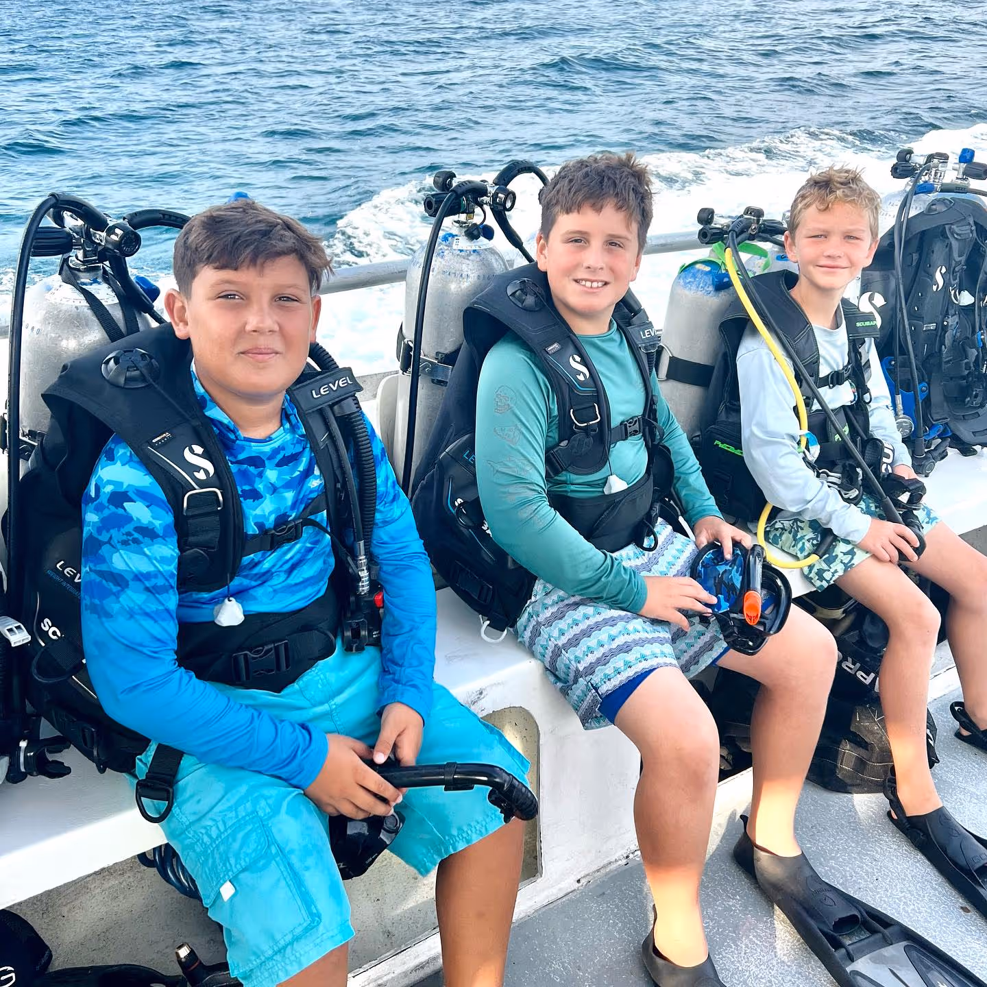 Three boys wearing scuba diving gear sitting on a boat with ocean water in the background.