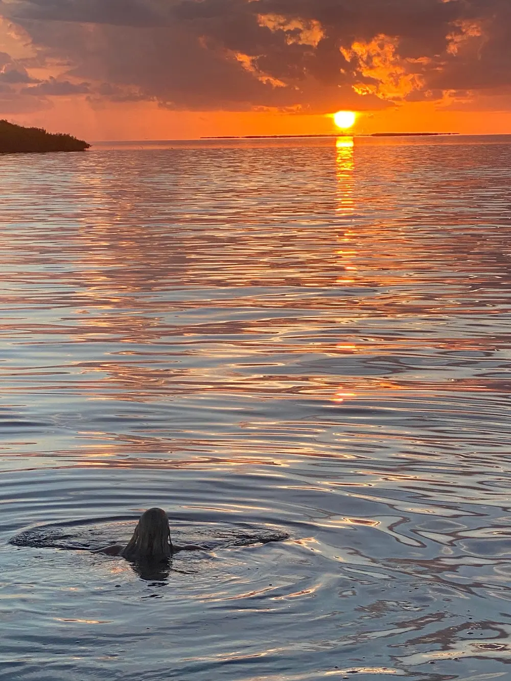 Person swimming in calm water reflecting an orange sunset with dark clouds.