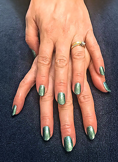 Two hands with green metallic nail polish, one wearing a gold ring with a small diamond, resting on a dark fabric surface.
