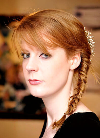 Portrait of a fair-skinned woman with red hair styled in a side braid wearing a black top.